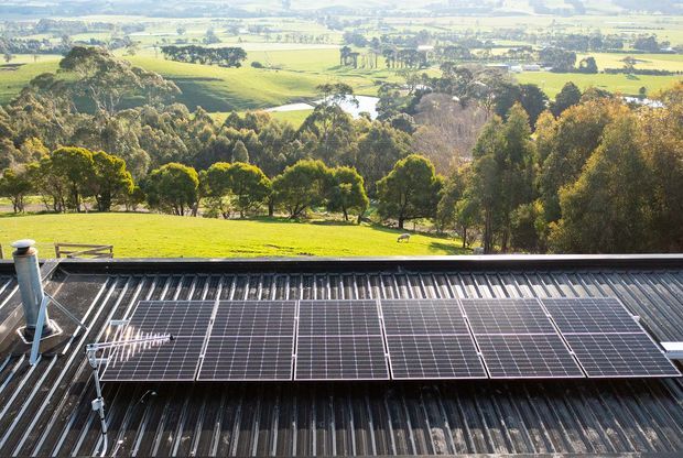 A rooftop solar photovoltaic array mounted on a corrugated metal roof, with a broad green rural landscape and patchwork farmland visible beyond. The image focuses on renewable-energy infrastructure and the surrounding pastoral setting.