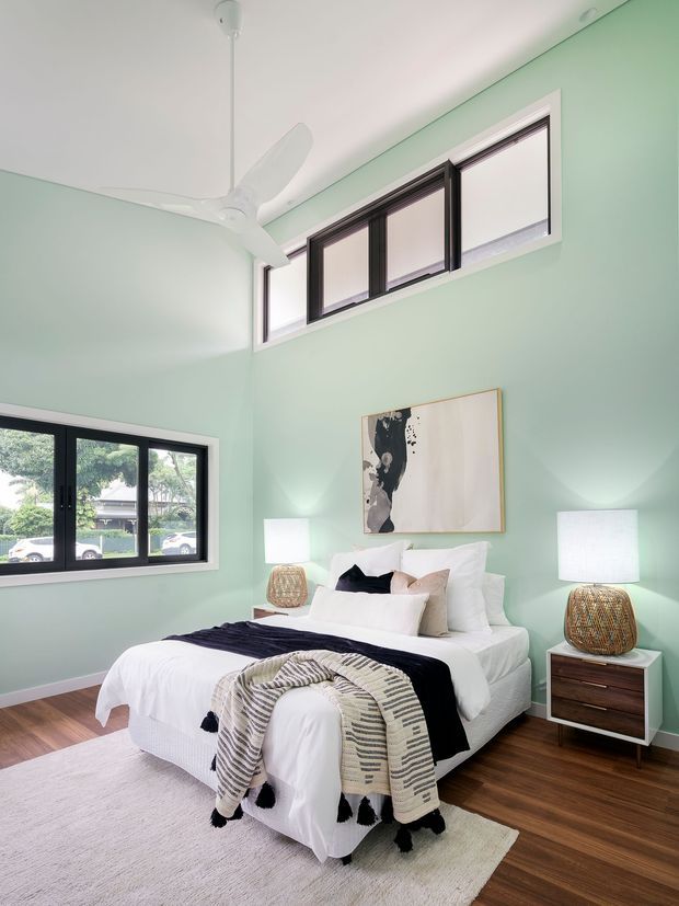 Bright, high-ceiling bedroom with pale mint-green walls, dark-framed windows, a white ceiling fan, and warm timber flooring. The room contains a neatly made white bed layered with black and neutral cushions and throws, matching bedside tables with woven lamps, abstract wall art, and a large pale rug. The upper clerestory windows and generous proportions create an airy feel.
