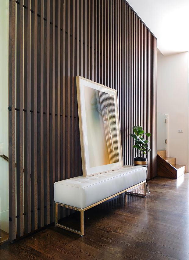 A refined modern interior entry hall with a dramatic full-height dark timber slatted feature wall. A white upholstered bench with a slim metallic base sits against the wall beneath a large framed artwork, accompanied by a potted plant. Dark timber flooring, a bright white ceiling, and a glimpse of a stairway create a warm, high-contrast composition.