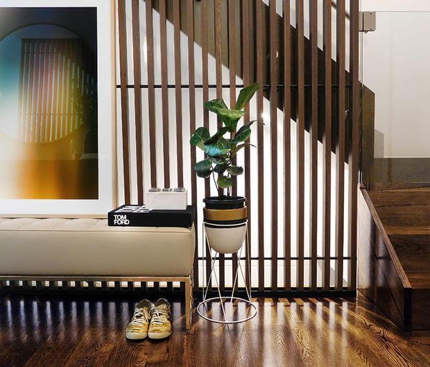 A refined interior entry or living area with a dark polished timber floor, a cream upholstered bench, a tall indoor plant, framed artwork, and a dramatic vertical timber slat screen beside a staircase. The slatted partition creates strong linear shadows and partially separates the seating area from the stairway. The space has a warm, contemporary mid-century-inspired character with high-contrast architectural detailing.