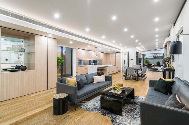 Open-plan contemporary living, dining and kitchen area with warm timber flooring, extensive built-in timber cabinetry, a central kitchen island, grey sofas, a black coffee table, and a dining setting. Recessed ceiling lights and large glazed openings create a bright, spacious interior with a view toward an outdoor area at the rear.