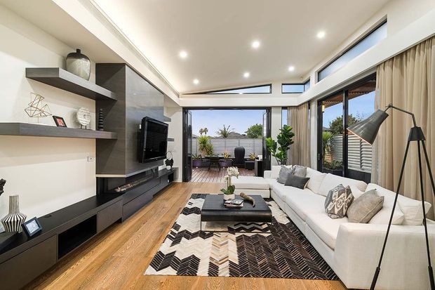 Bright contemporary living room with warm timber flooring, a large white sectional sofa, black built-in entertainment joinery and open shelving, a textured patterned rug, and floor-to-ceiling glazing opening to an outdoor terrace. High-level clerestory windows and recessed ceiling lights enhance the airy feel.