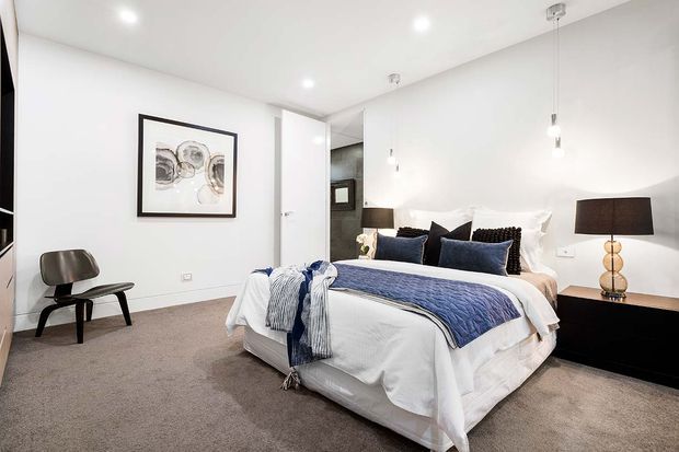Bright, contemporary bedroom with a large upholstered bed dressed in white bedding and a blue quilt, dark blue accent cushions, a black bedside cabinet, and a sculptural table lamp. The room has white walls, recessed ceiling lights, pendant lights, soft grey carpet, framed abstract artwork, and a dark lounge chair. A partially visible doorway and dark tiled area sit behind the bed.