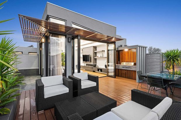 Contemporary rooftop terrace and outdoor living area with timber decking, black wicker seating, a glass dining table, and lush potted planting. A timber pergola shades the open living space, which connects directly to a bright modern interior through large glazed doors. A compact outdoor kitchen or bar with vertical timber screening is visible beside the terrace.