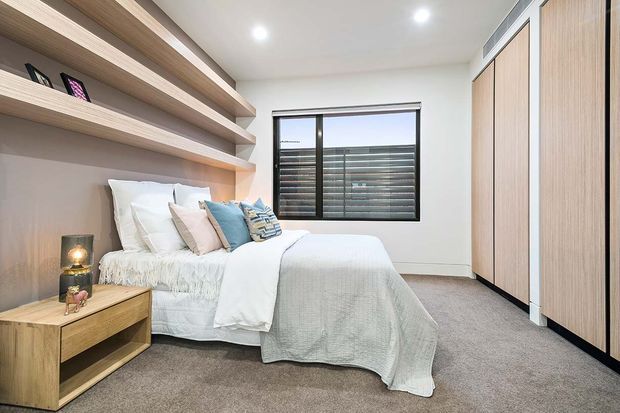 A compact contemporary bedroom with a neatly made bed, built-in timber-look open shelving and wardrobe cabinetry, a timber bedside table, and a large black-framed window. Soft neutral finishes, layered bedding, and recessed ceiling lights create a warm, calm interior.
