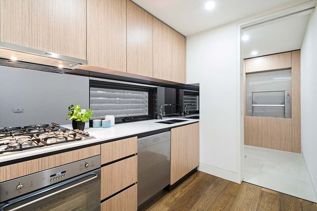 Compact contemporary kitchen with light timber-grain cabinetry, dark accents, a white benchtop, integrated stainless-steel appliances, gas cooktop, sink, and a window with layered blinds. The kitchen opens to a light-tiled utility or pantry area, while warm wood-look flooring contrasts with the white walls.