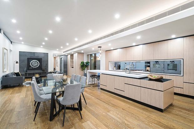 A bright, open-plan contemporary kitchen and dining area with extensive light timber cabinetry, a large central island, integrated appliances, and a dining table with upholstered chairs. A living area with a dark feature wall and television is visible in the background. Recessed ceiling lighting and linear air conditioning reinforce the clean modern fit-out.