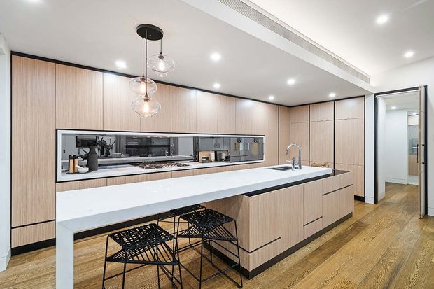 Contemporary open-plan kitchen with extensive light timber cabinetry, a long white island bench with integrated sink and seating, mirrored splashback, black wire bar stools, pendant lights, and warm timber flooring. The space has clean lines, recessed lighting, and a bright, refined finish.