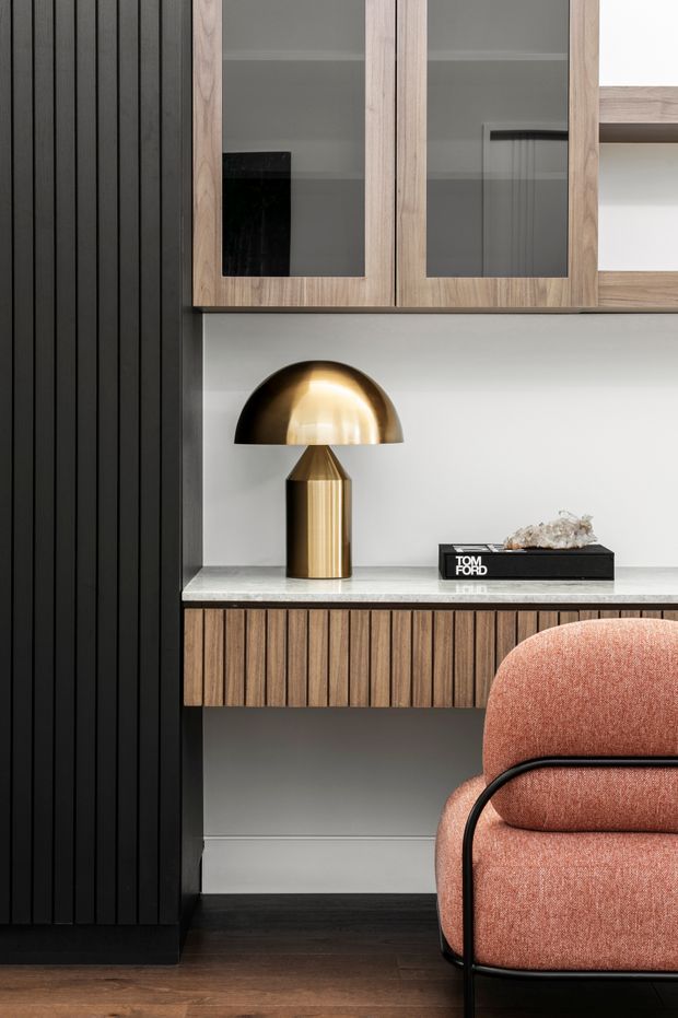 Close-up of a refined built-in study or home office joinery arrangement. The space features dark vertical wall panelling, a timber-framed glass-fronted overhead cabinet, a light stone benchtop, fluted timber cabinetry below, and a sculptural brass table lamp. A blush upholstered armchair with a black metal frame occupies the foreground. The palette is warm and sophisticated, combining black, timber, brass, stone, and muted coral tones.