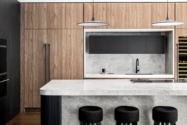 Contemporary kitchen featuring a large pale stone island with three black upholstered stools, integrated dark cabinetry, warm vertical timber cabinetry, a matching stone splashback and black pendant lights. A built-in wine storage unit is visible at the right.