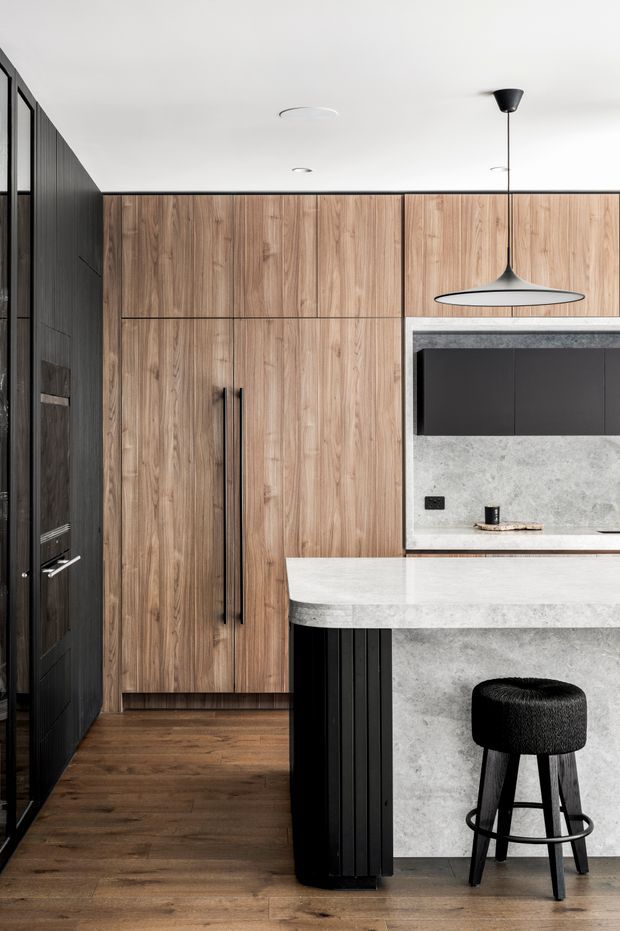 A contemporary kitchen with full-height warm timber cabinetry, a dark charcoal wall of joinery, a pale stone island and matching stone splashback. The island includes black vertical fluted detailing and seating for one, complemented by a black upholstered bar stool and a minimalist pendant light.