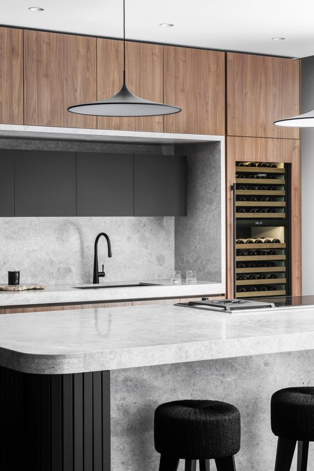 Contemporary kitchen with a large pale stone island and integrated cooktop, extensive warm timber cabinetry, dark grey overhead cupboards, concrete-look wall finishes, a black gooseneck tap, and built-in wine storage. Black pendant lights and upholstered bar stools add strong contrast.
