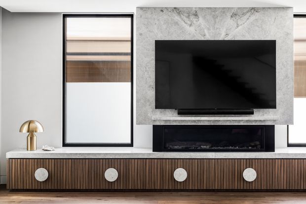 Contemporary living room media wall featuring a large black television mounted on a pale grey veined stone slab, a linear black fireplace below, and a long ribbed dark timber entertainment cabinet with oversized circular pulls. Black-framed windows with textured blinds and a sculptural brass table lamp add contrast. The room has a refined, monochromatic material palette with warm timber detailing.