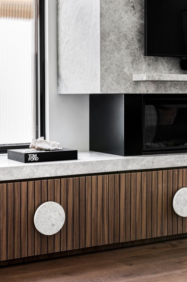 Close-up of a sophisticated living room media wall featuring a black built-in fireplace and television, pale grey stone cladding and mantel, dark vertical timber-slat cabinetry with large round stone handles, and dark timber flooring. A tall window with vertical blinds is visible at left.