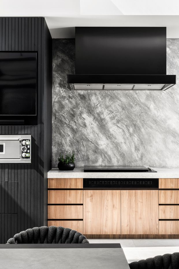 Contemporary kitchen with a dramatic black rangehood, dark vertically ribbed wall cabinetry and integrated appliances, contrasted with warm timber cabinetry and a mottled grey stone-look splashback. A pale stone or concrete island occupies the foreground, with dark upholstered seating visible at the lower edge.