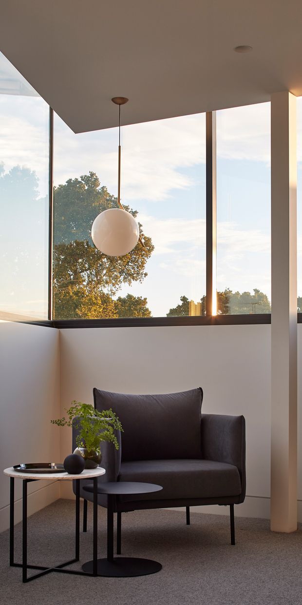 A refined, minimalist sitting corner with a dark upholstered armchair, two small black metal side tables, a potted plant, and a spherical pendant light. Large floor-to-ceiling windows provide expansive views of trees and warm late-day light. The space has a white plaster ceiling and walls, slim dark window frames, a carpeted floor, and a clean contemporary architectural composition.