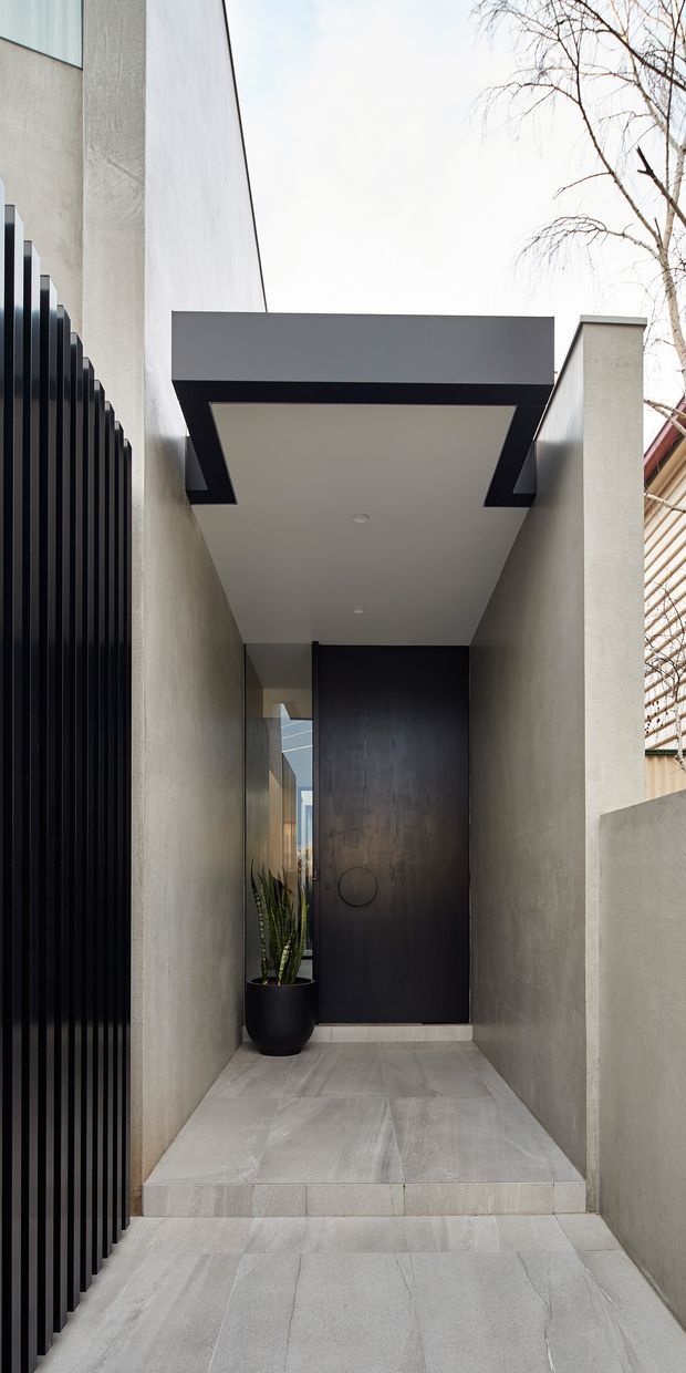 A minimalist contemporary residential entry passage viewed from outdoors, with pale grey rendered walls, large-format light grey tiled paving, a tall dark timber or dark-finished front door, black vertical metal screening along the left boundary, and a projecting flat canopy with black trim. A potted plant sits beside the entrance, while neighbouring residential fabric and a leafless tree are visible beyond.