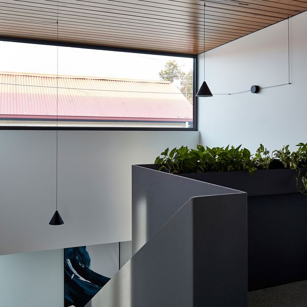 A minimalist interior stairwell or landing with a sculptural dark geometric stair balustrade, white walls, a planter integrated along the upper edge, and slim black pendant lights. A large horizontal window provides views of a nearby corrugated metal roof, while the ceiling is lined with warm timber battens. The space has clean contemporary detailing and strong geometric forms.