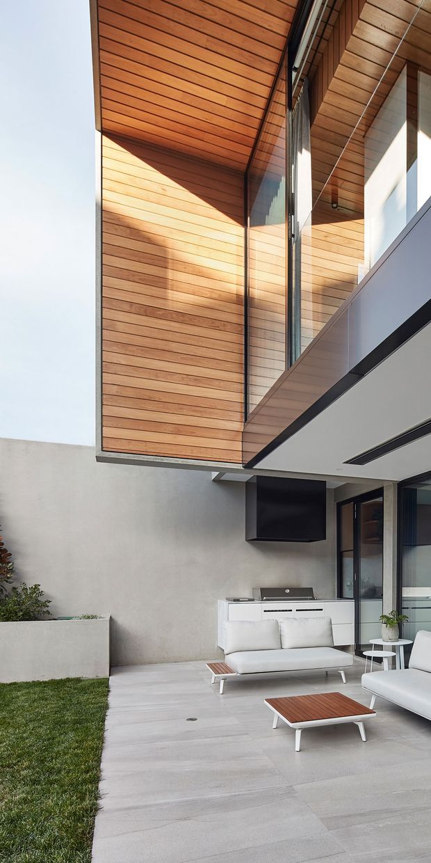 A contemporary courtyard patio beneath a dramatic cantilevered upper level. Warm timber lining covers the soffit and upper wall, contrasted with smooth pale rendered walls, dark metal framing, extensive glazing, and a clean concrete-paver terrace. The outdoor lounge includes white seating and low timber-topped tables, with a built-in white barbecue kitchen against the wall. A narrow grass strip and raised planter add greenery.