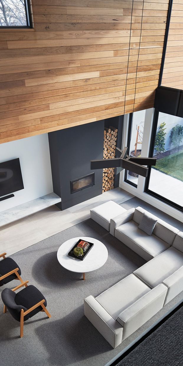 A double-height contemporary living room viewed from above, featuring extensive warm timber wall and ceiling lining, a dark charcoal fireplace wall with built-in firebox and stacked firewood, expansive black-framed glazing, and a large pale sectional sofa arranged around a round white coffee table. A sculptural dark ceiling fan hangs over the seating area, with a television and armchairs completing the room. The space opens visually toward a small courtyard garden.