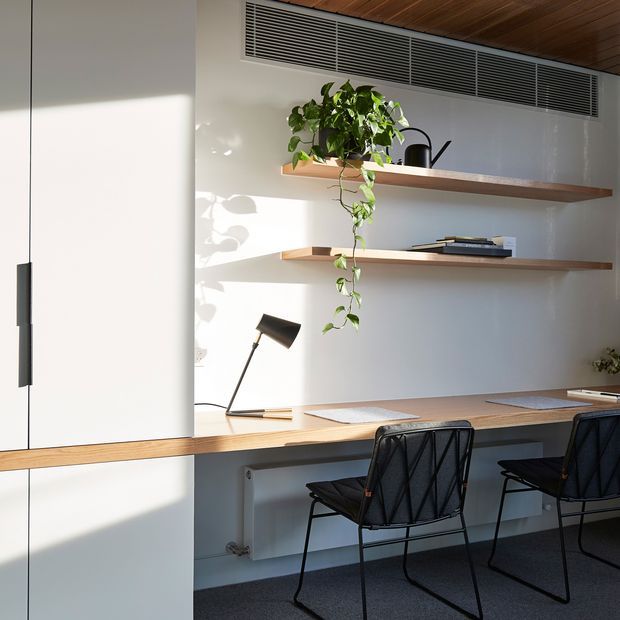 A bright, minimalist built-in home workspace with a long timber desk, open timber shelves, black task lamp, two black wire-frame chairs, concealed cabinetry, and a trailing indoor plant. White walls and cabinetry contrast with the warm timber ceiling and joinery, while linear air-conditioning grilles run along the upper wall.