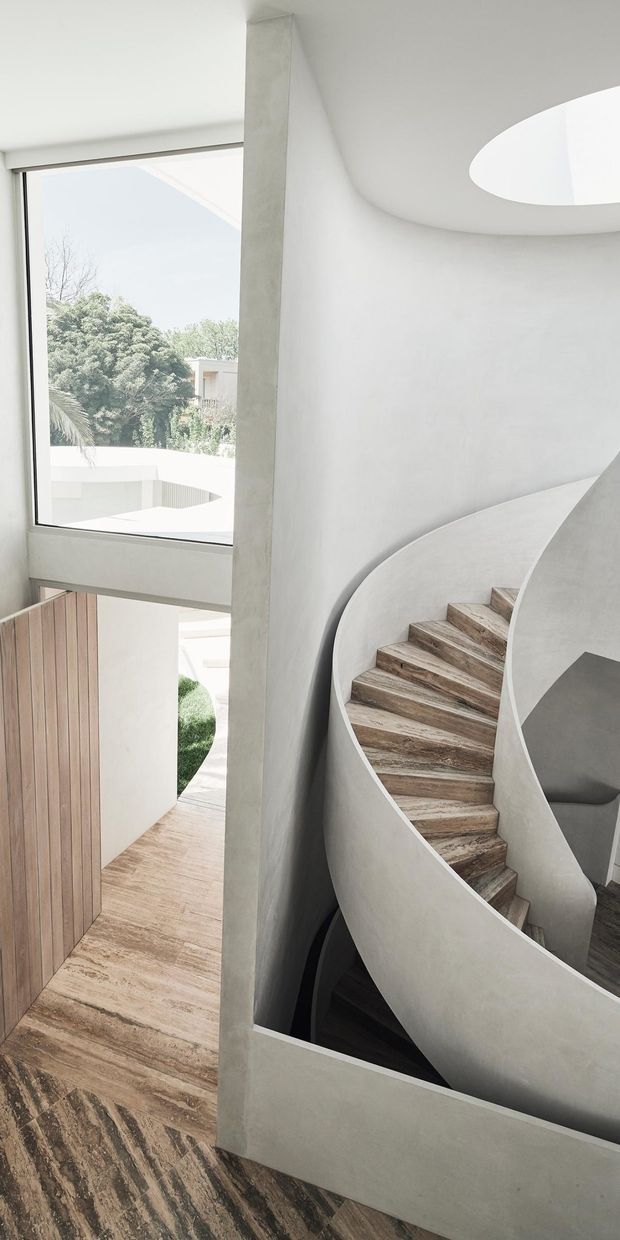 A sculptural interior stairway with a sweeping curved plaster or concrete balustrade and matching rounded wall. The staircase has richly textured timber or stone-look treads, while a large window brings in views of greenery and daylight. A circular skylight or roof opening illuminates the upper landing, and a timber-lined doorway is visible below. The overall composition is minimalist, monolithic, and highly organic in form.