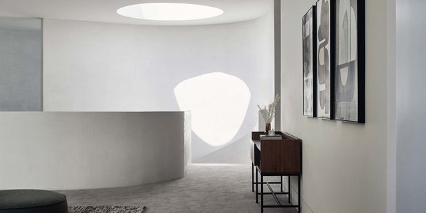 A minimalist interior hallway or landing with smooth white curved walls, a low partition wall, grey carpeted flooring, and a sculptural circular ceiling opening or light feature. A dark timber console with slender black metal legs sits against the right wall beneath vertically oriented monochrome artwork. The space is sparsely furnished and strongly geometric, with soft natural light creating a rounded highlight on the rear wall.