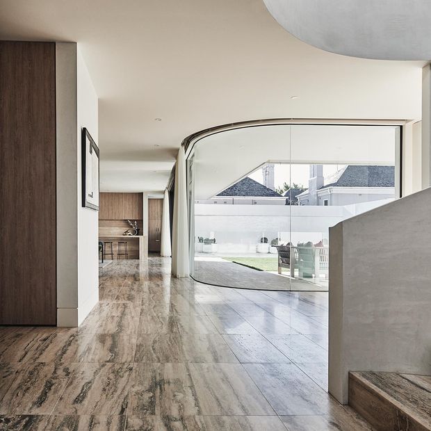 A refined contemporary interior hallway or entry space with a dramatic curved floor-to-ceiling glazed wall opening onto a bright courtyard. The space features large-format veined stone-look flooring, dark timber built-in cabinetry, smooth warm-white walls and ceiling, recessed downlights, and a sculptural curved ceiling detail. A partial stair or raised platform with dark timber trim is visible in the foreground.
