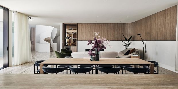 Open-plan contemporary living and dining area with a long timber dining table and dark upholstered chairs in the foreground, a pale sofa and lounge seating beyond, and a dramatic curved white staircase at the rear. The room features a full-height warm timber feature wall with integrated shelving and cabinetry, pale stone or plaster lower walls, light timber flooring, floor-to-ceiling glazing with sheer curtains, recessed ceiling lighting, and restrained sculptural decor.