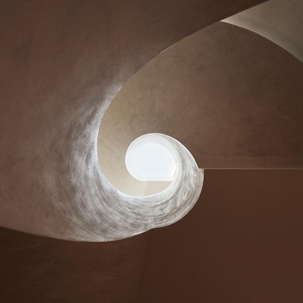 Upward-looking view of a sculptural spiral stairway or curved architectural ceiling enclosure, finished in warm brown plaster. A circular skylight or roof opening at the centre brings in bright natural light, emphasizing the sweeping organic geometry and textured surface.