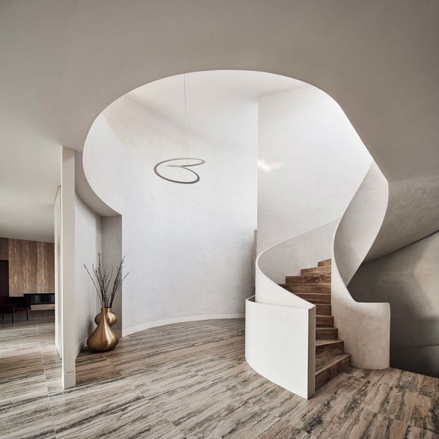 A sculptural interior stairway with a sweeping curved white plaster balustrade and matching curved ceiling void. Timber-toned stair treads rise through the centre, while pale stone- or timber-look flooring extends across the open foyer. A minimalist pendant light, simple decorative vase, and glimpses of dark timber cabinetry are visible to the left. The space is bright, refined, and highly contemporary, with strong organic curves and a clean neutral palette.