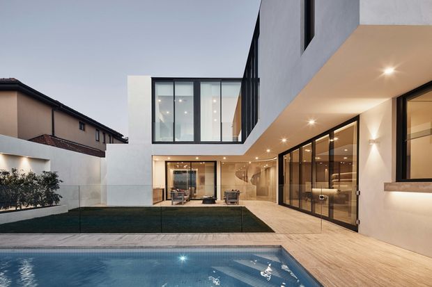 Contemporary multi-level residential house viewed from the rear, with a white rendered facade, extensive black-framed glazing, deep covered outdoor walkway, timber-toned decking, landscaped courtyard lawn, and a swimming pool in the foreground. Warm recessed exterior lighting illuminates the ground-floor indoor-outdoor living areas, while a double-height glazed volume projects above the courtyard.