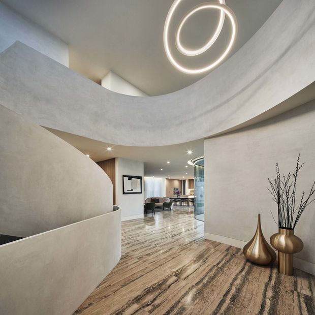 A refined contemporary residential interior showing a sweeping curved staircase or balustrade beneath a sculptural circular pendant light. The open-plan space extends toward a dining and living area, with pale textured plaster walls, curved architectural forms, recessed lighting, and dramatic veined stone flooring. Metallic decorative vessels with branches sit against the wall in the foreground.