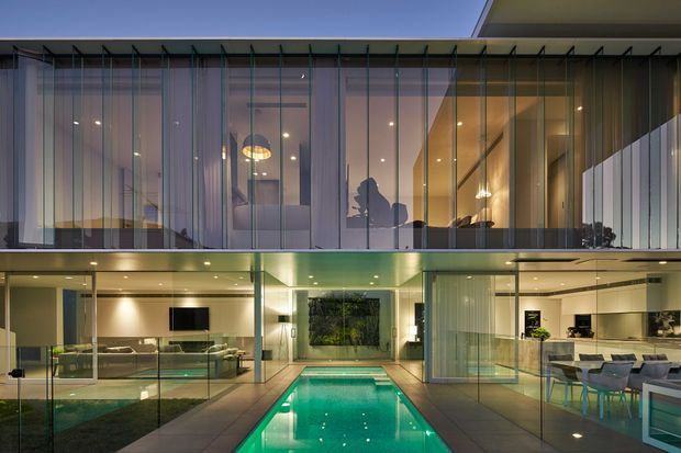 Twilight view of a contemporary multi-level residence organized around a narrow illuminated swimming pool courtyard. Extensive floor-to-ceiling glazing, slender vertical framing, projecting roof planes, and transparent balustrades create a strongly indoor-outdoor connection. The lower level reveals a living area and open kitchen/dining space, while the upper level contains glazed rooms or a balcony zone. The palette is predominantly neutral grey, beige, glass, and warm timber-toned lighting, with the pool providing a vivid turquoise accent.