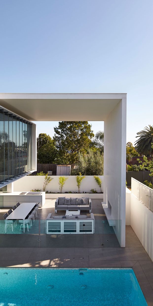 A high-angle view of a contemporary residential outdoor entertaining area with a swimming pool in the foreground. The space features a large white flat-roofed architectural frame, extensive floor-to-ceiling glazing, a dining setting, outdoor lounge seating, dark tiled paving, rendered walls, and landscaped planting. The design is bright, open, and strongly connected to the pool courtyard.