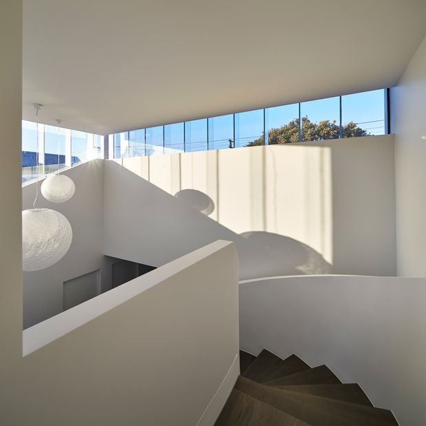 A minimalist interior stairway with dark timber treads, white rendered walls, and a sculptural curved balustrade. High clerestory glazing brings in strong natural light, creating pronounced geometric shadows across the walls. Two large spherical pendant lights hang beside the stairwell, with glimpses of trees and neighbouring structures visible through the upper windows.