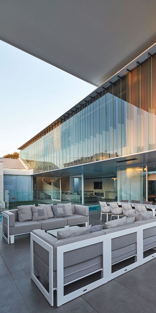 A contemporary outdoor entertaining terrace framed by a dramatic cantilevered roof and a two-storey glass facade. The space contains large pale-grey upholstered outdoor sofas, white-framed lounge furniture, and a dining setting overlooking a narrow pool or water feature. Floor-to-ceiling glazing reflects the surrounding landscape, while timber vertical screening and slender dark structural elements add warmth and rhythm to the facade.