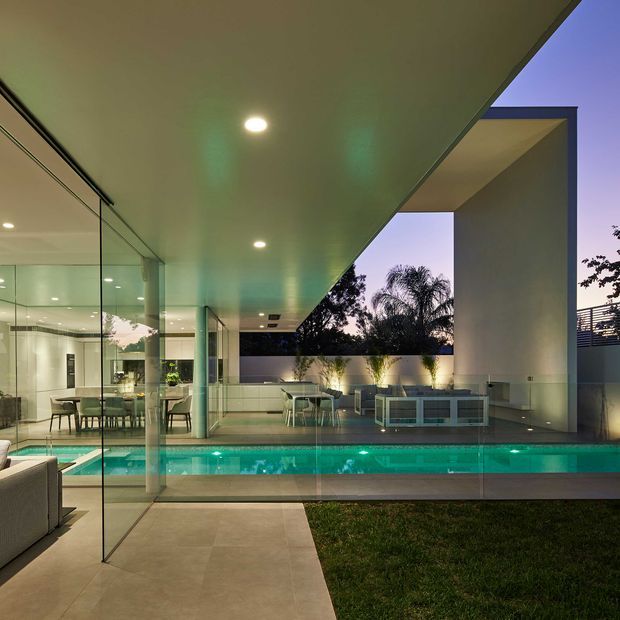 A dusk view of a sophisticated modern residence centred on a narrow illuminated swimming pool and covered outdoor terrace. Large frameless glass walls reveal a contemporary living and dining area, while a broad flat-roof canopy, recessed downlights, pale tiled paving, sculptural architectural columns, and built-in outdoor seating define the space. The composition emphasizes seamless indoor–outdoor connection and strong geometric lines.