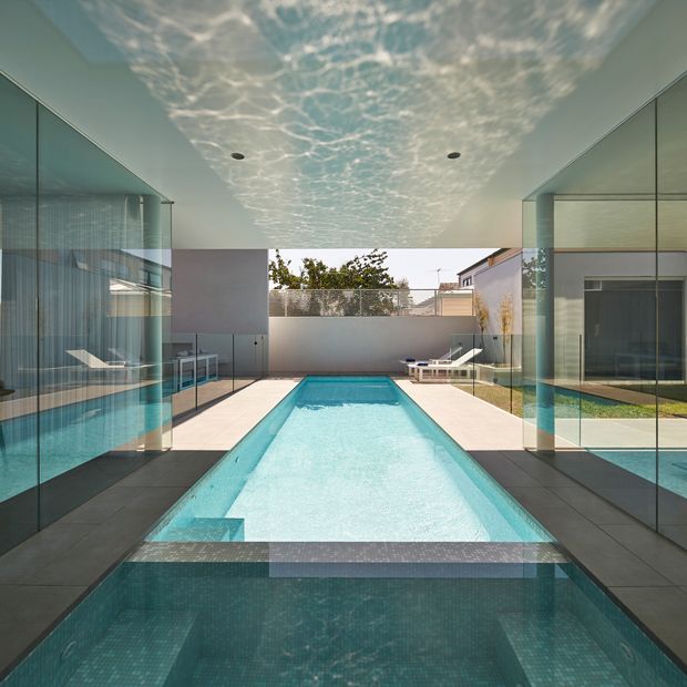 A modern indoor-outdoor pool pavilion with a long rectangular swimming pool extending through a glazed enclosure toward an open courtyard. Full-height glass walls and doors flank dark tiled flooring, while a white ceiling features a rippled, water-reflective effect with recessed downlights. Minimal white loungers and contemporary landscaping are visible beyond the pool.
