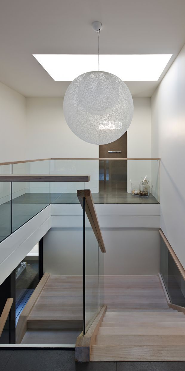 A contemporary multi-level stairwell and landing with pale timber stairs, clear glass balustrades, slim dark handrails, and white walls. A large textured spherical pendant hangs beneath a bright rectangular skylight, illuminating the open stair void and upper landing. A dark-toned doorway and simple wall light are visible at the rear.