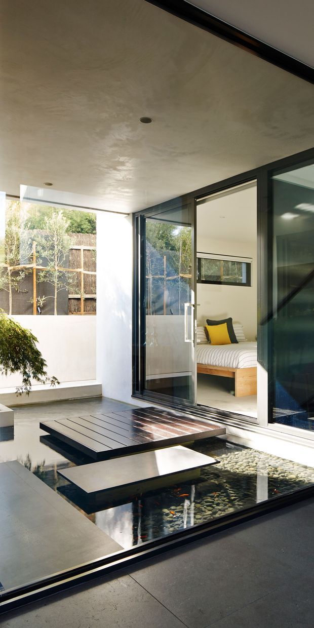A modern indoor-outdoor courtyard viewed from a covered living area, featuring dark-framed floor-to-ceiling glazing, a shallow reflective pond with pebble edging and stepping slabs, and a floating timber platform. A compact bedroom with a striped bed and yellow accent cushion is visible through the open glass doors. The space has a smooth warm-toned ceiling, pale walls, and strong connections to a planted courtyard.