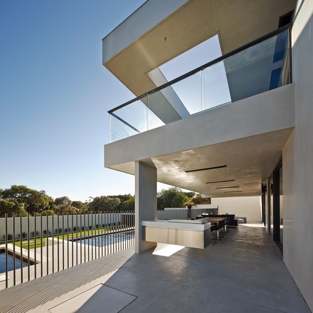 Modern multi-level residential exterior with a covered outdoor entertaining area overlooking a swimming pool. The space features a large built-in outdoor kitchen or bar with seating, smooth rendered concrete surfaces, slim dark-framed glazing, clear glass balustrades, and a geometric roof opening that brings in sunlight. A vertical metal fence encloses the pool area.