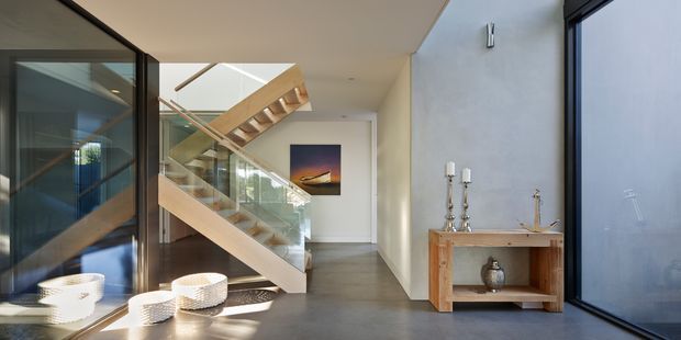 Contemporary interior hallway and stairway with a floating timber staircase, clear glass balustrades, dark-framed glazing, polished grey concrete flooring, and pale plaster walls. A timber console with decorative objects sits against the wall, while strong daylight enters through the glazed stair and adjacent windows.