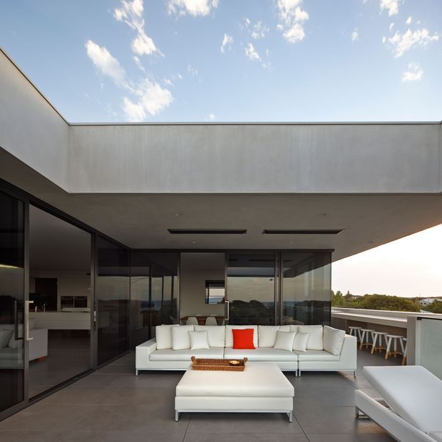 A contemporary rooftop or upper-level outdoor lounge beneath a broad flat concrete canopy. The space features a large white sectional sofa, matching ottoman, and a white lounge chair arranged on large grey floor tiles. Full-height dark-framed glazing connects the lounge to an interior kitchen or living area, while a glass balustrade opens toward a leafy suburban view. The composition is minimalist, spacious, and refined, with a vivid red cushion as a small accent.