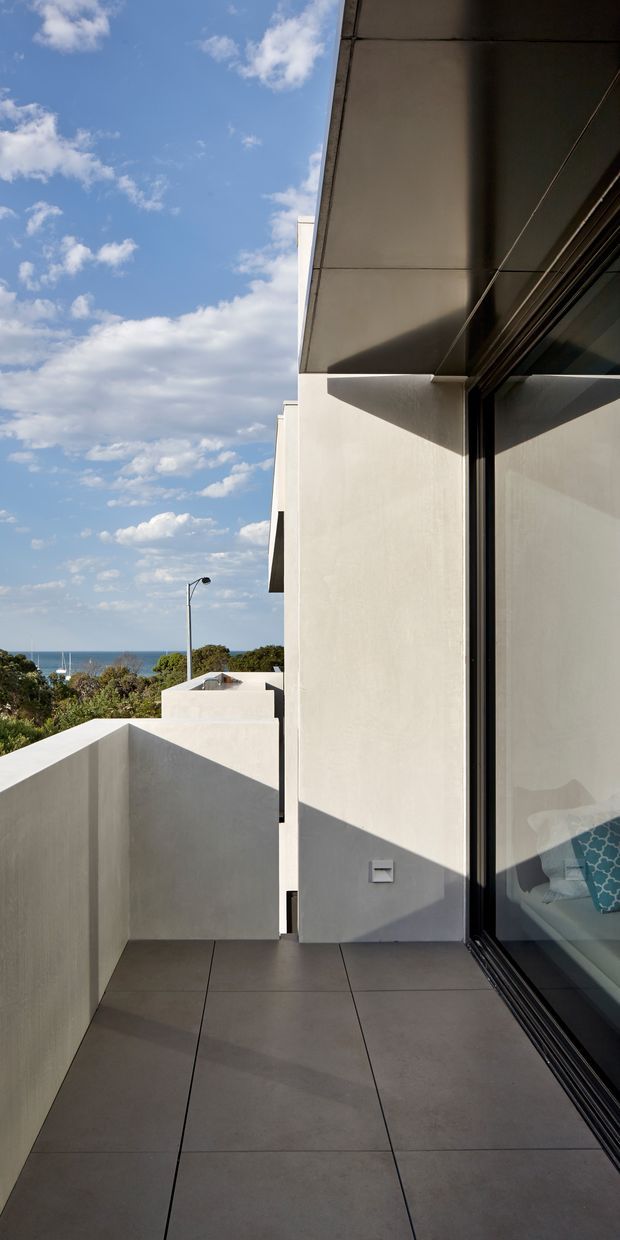 A narrow elevated outdoor terrace or balcony beside a modern white rendered building. Large grey square-format tiles cover the floor, with a solid rendered privacy wall and a broad projecting roof or eave above. Full-height dark-framed sliding glazing reflects the exterior and reveals part of an interior room with cushions. The terrace overlooks trees and a visible strip of blue ocean beneath a bright, partly cloudy sky. Strong geometric shadows and clean modern lines dominate the composition.