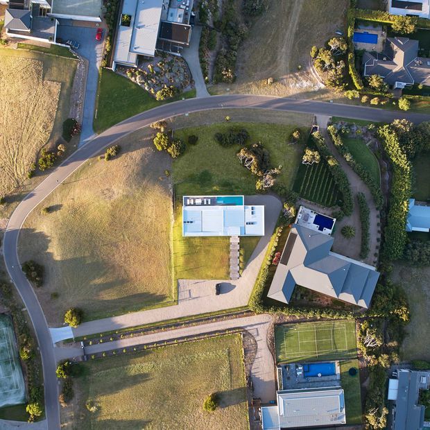 Aerial drone photograph looking straight down over a suburban residential neighbourhood. Detached homes with varied roof forms, landscaped gardens, driveways, open grassy lots, curving paved roads, and a fenced tennis court with a swimming pool are visible.