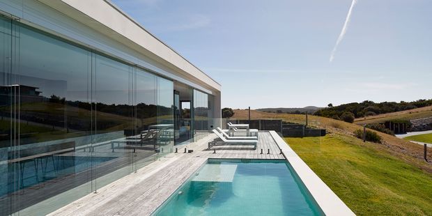 Exterior view of a minimalist contemporary residence set in a rolling rural landscape. The image shows a long, low white facade with extensive floor-to-ceiling glazing, a timber-toned deck, and a narrow rectangular swimming pool adjoining the building. Outdoor lounge furniture sits beside the pool, with open lawn and distant hills beyond.