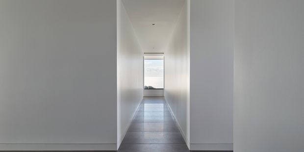 A long, narrow minimalist hallway with smooth white walls, a plain white ceiling with recessed lighting, and large-format grey tiled flooring. The corridor leads toward a bright window with a partially lowered blind and a distant exterior view, creating a strong linear perspective.