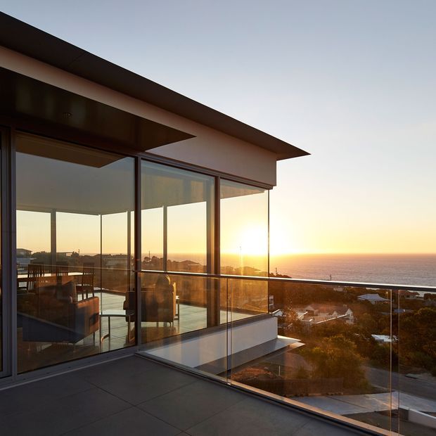 Modern coastal residence at sunset, viewed from a spacious balcony or terrace with frameless glass balustrades. Floor-to-ceiling glazing reveals an open-plan interior with a dining table and lounge seating, while the projecting flat roof and clean geometric lines frame expansive ocean views.