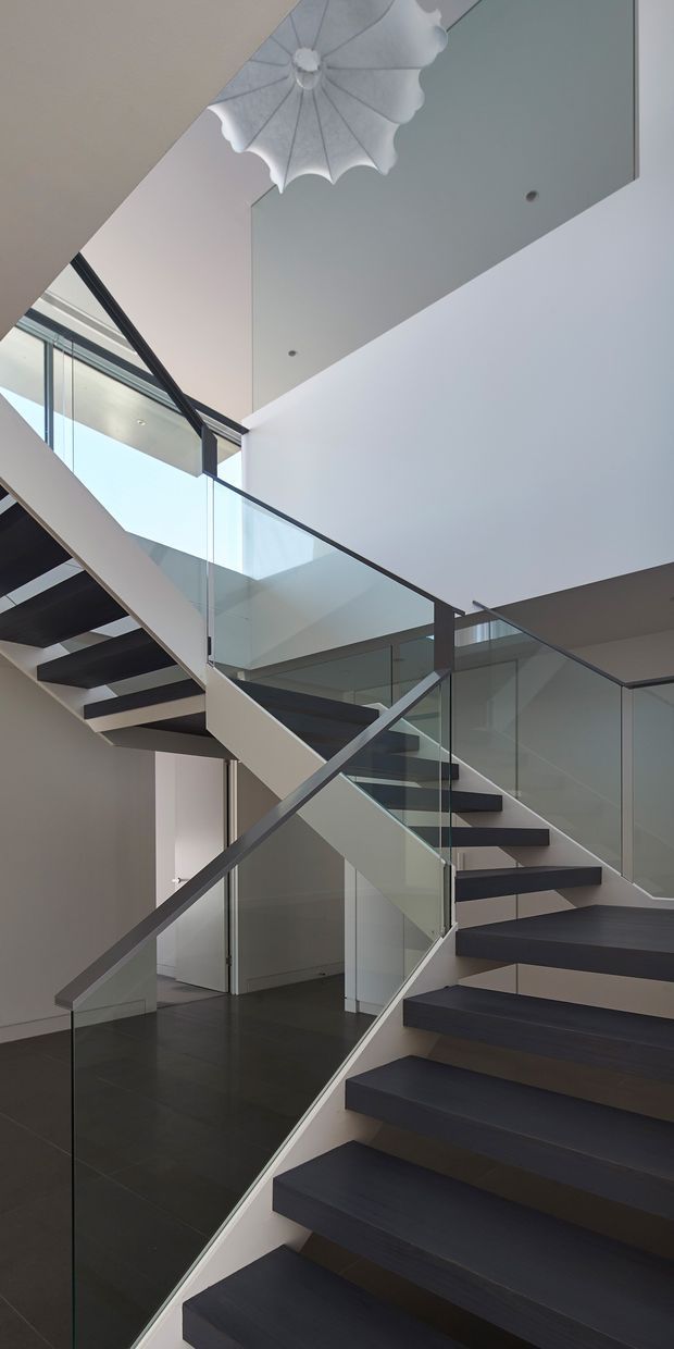 A contemporary interior stairway viewed upward through a multi-level stair hall. Dark floating timber treads contrast with white structural walls and floors, while clear glass balustrades and slim dark metal handrails create a light, open composition. Large glazing brings in daylight, and a sculptural white pendant light hangs near the ceiling. The space has a strong vertical sense of height and a minimalist architectural character.
